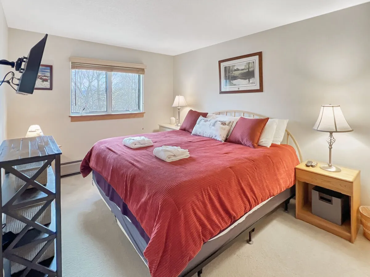 A cozy bedroom featuring a bed with a red comforter, wall-mounted television, nightstands with lamps, and a window letting in natural light. The space exudes relaxation and privacy, ideal for unwinding after an active day.