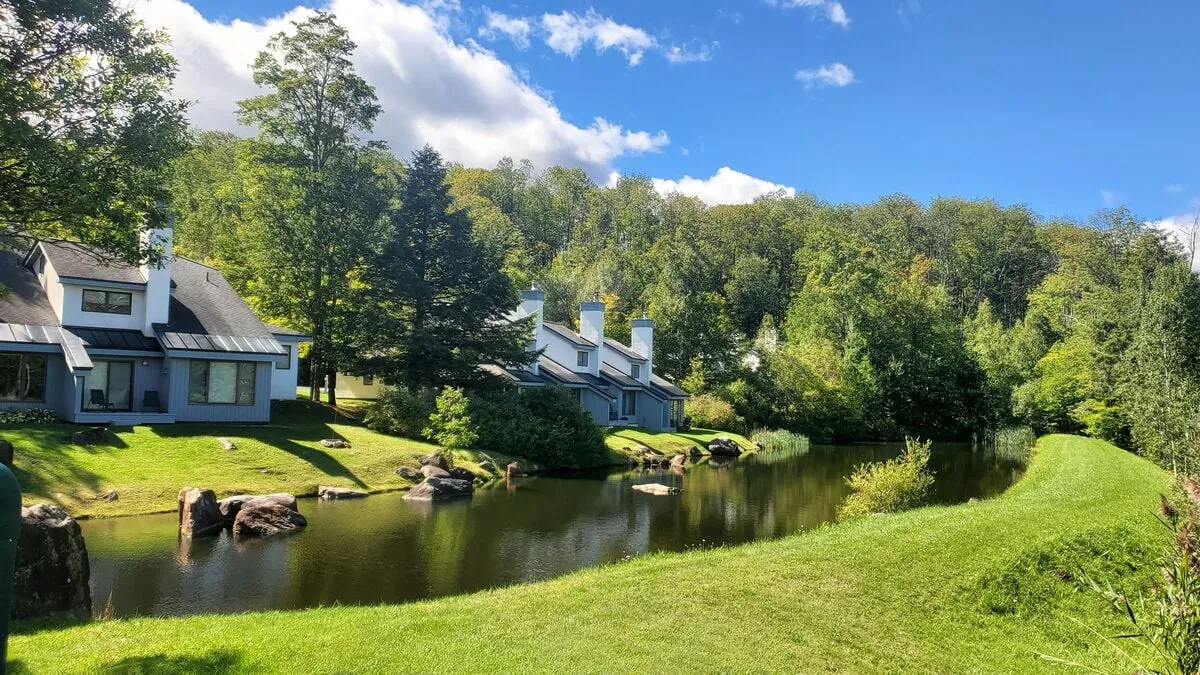 This tranquil exterior image features houses overlooking a serene pond, surrounded by trees and a well-kept lawn. The setting captures the natural beauty of Killington, inviting outdoor activities like walking, relaxing by the water, or enjoying sunset views.