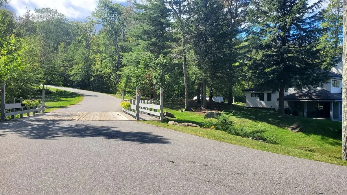 A peaceful exterior view of a winding road, wooden bridge, and lush greenery, leading toward a house in the background. The scene hints at scenic drives, romantic walks, and the serene natural environment that defines the retreat.