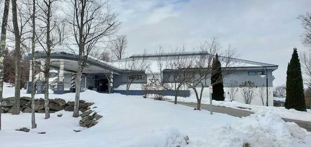 A modern condo building is surrounded by a pristine, snow-covered landscape and bordered by a line of tall trees. The view highlights the main entranceway, with inviting architecture that hints at warmth and comfort inside. The snowy setting captures the spirit of winter sports and après-ski relaxation, while the building’s design suggests access to upscale amenities.