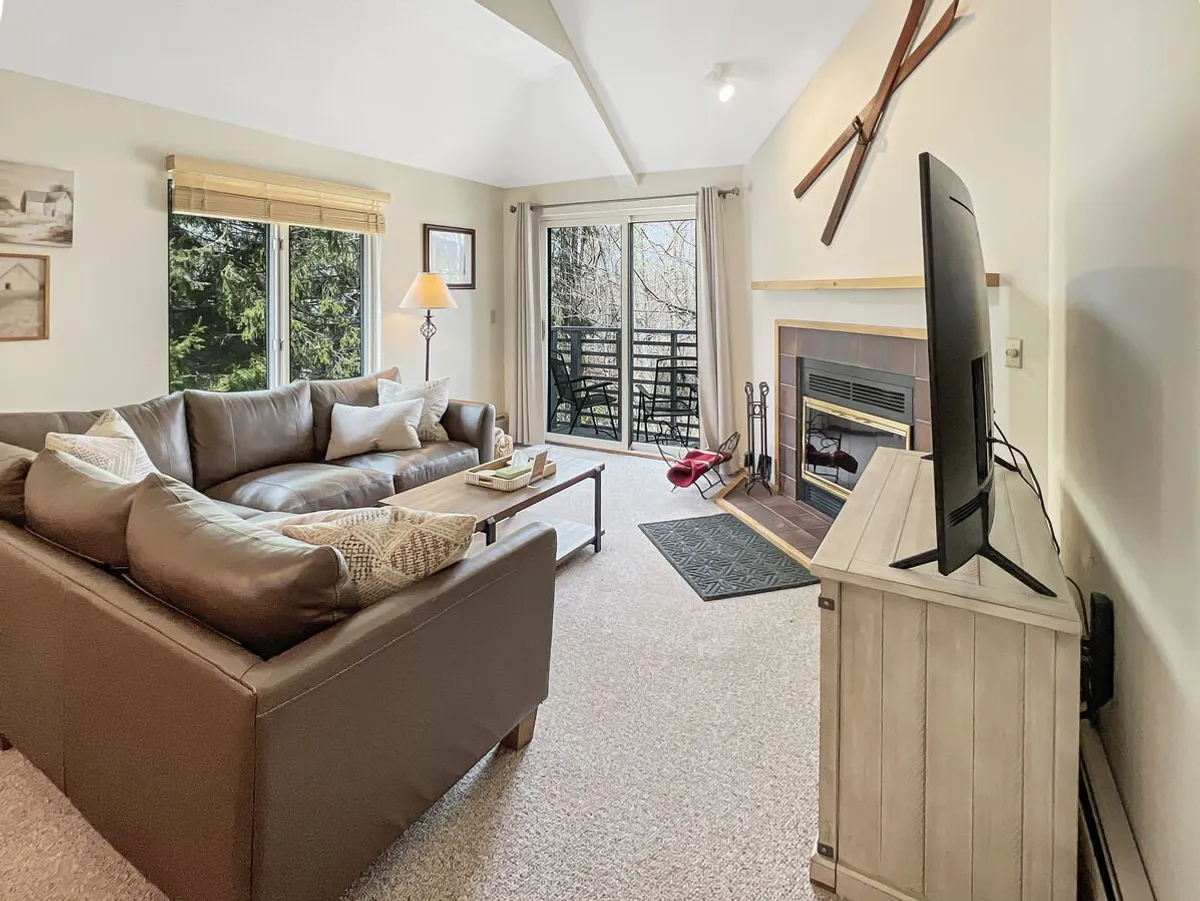 A warm open-plan living area featuring a plush sofa facing a wood-burning fireplace, with a television mounted nearby and a sliding glass door letting in natural light. The scene exudes comfort and is clearly intended for relaxation, entertaining, or gathering with family after a day outside.