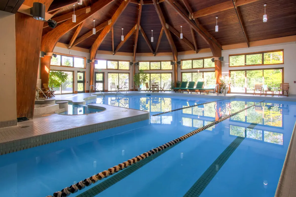 This image showcases a serene indoor pool area featuring a sparkling swimming pool, a bubbling hot tub, large windows allowing natural light to flood in, wooden beams for a cozy touch, and comfortable lounge chairs positioned around the water. The inviting setup hints at both relaxation and socializing opportunities in a revitalizing environment.