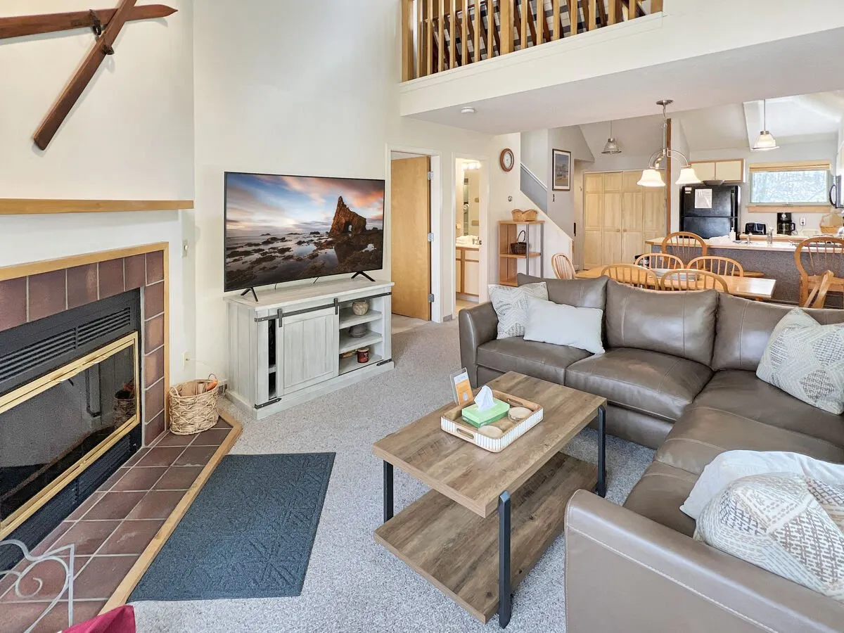 The image showcases a warm, inviting living space featuring a corner sofa facing a crackling fireplace, a flat-screen television above the mantle, and an adjacent dining area perfect for gathering. The space glows with soft lighting and rich wood accents, evoking comfort and relaxation.